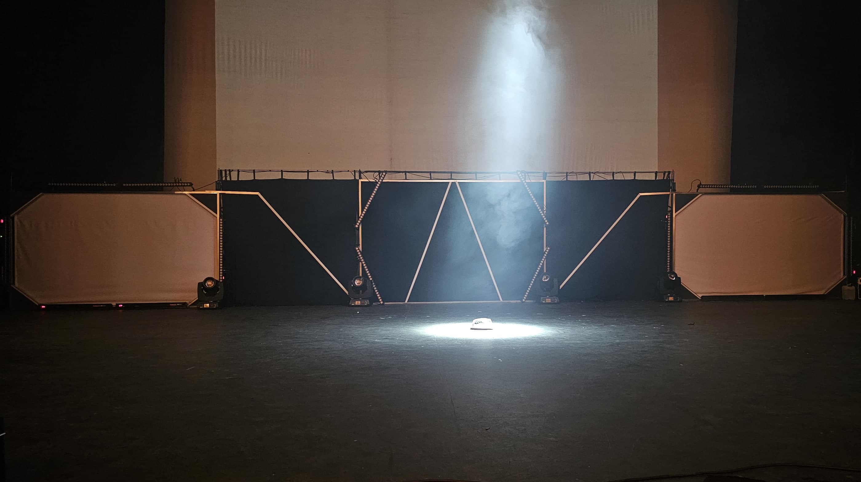 La escenografía montada en el escenario, de frente: una pared de paneles con líneas de luz en zigzag, cañones de luz y humo.